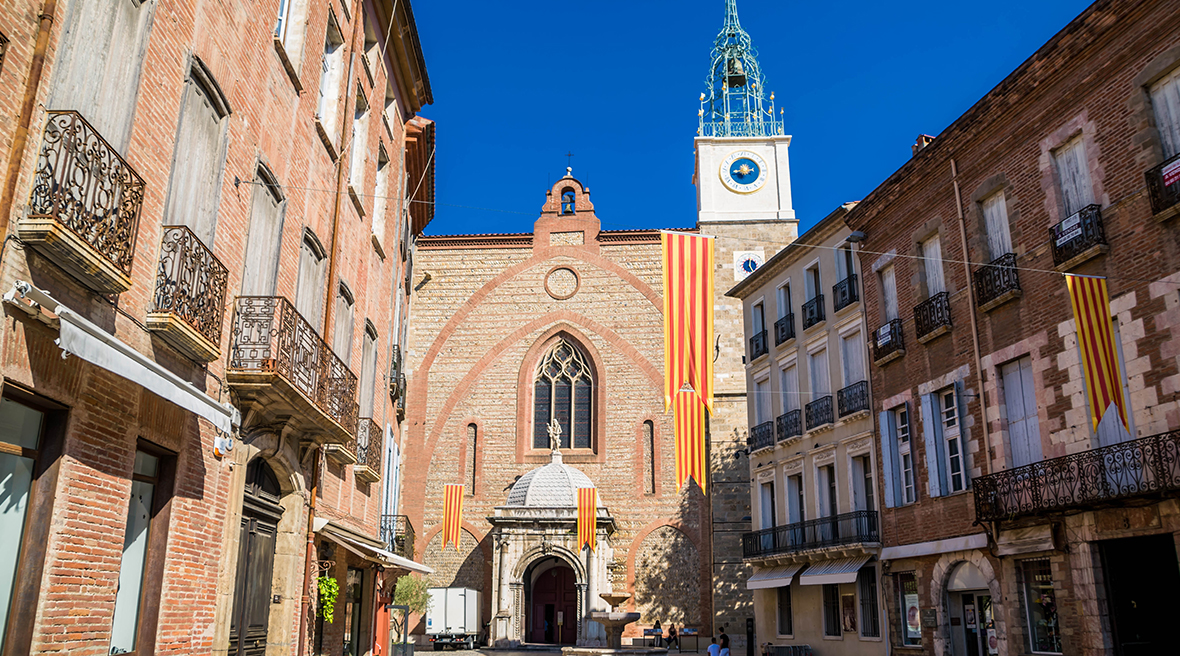 City square in Perpignan with red brick buildings, balconies, and the cathedral spire in the background
